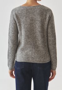 Osoba w szarym swetrze z dzianiny i ciemnoniebieskich dżinsach, stojąca tyłem do aparatu na jednolitym tle.