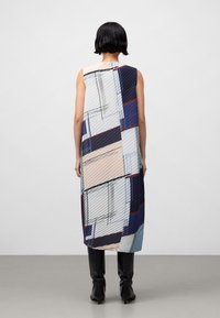 Vrouw met kort zwart haar draagt een mouwloze jurk van knielengte met een abstract geometrisch patroon en zwarte laarzen tot halverwege de kuit, staande op een grijze vloer.