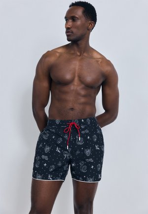 Mišičast moški, oblečen v črne kopalne hlače z belimi vzorci in rdečo vrvico, stoji z rokami za hrbtom in gleda v stran.