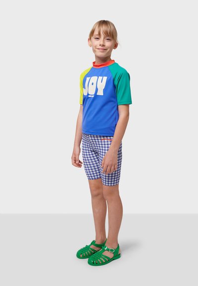 Färgglad kortärmad rash guard i blått, grönt och gult med texten "JOY", tillsammans med blå rutiga shorts och gröna sandaler.