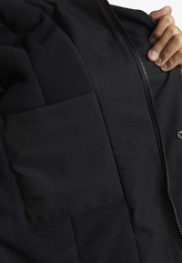 Sort jakke med et blødt, tekstureret fleece-sektion og en glat stoflomme. Lynlås med en robust zip-slider.