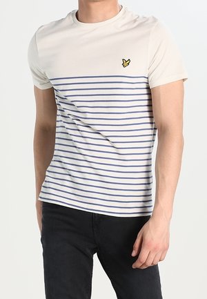 Mand iført en cremefarvet t-shirt med blå vandrette striber og et lille gult fuglelogo på brystet, kombineret med sorte bukser.