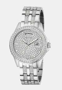 Zegarek Guess z srebrną bransoletą ze stali nierdzewnej, okrągłą tarczą, ozdobioną cyrkoniami oraz teksturowanym cyferblatem. Posiada okienko daty oraz wskazanie dnia.
