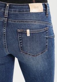 Jeans de mezclilla azul con un corte ajustado, cintura de altura media y un pequeño bolsillo trasero con etiqueta. La tela tiene un acabado ligeramente texturizado.