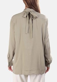 Blusa beige chiaro con maniche lunghe, dettaglio del fiocco sulla schiena e tessuto morbido. Presenta una chiusura con bottoni e una vestibilità rilassata.