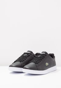 Svarta lädersneakers med rund tå, vit gummisul, matchande snörning och grön Lacoste-logotyp på sidan. Slät textur med minimal sömnad.