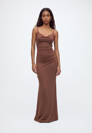 Femme aux longs cheveux foncés portant une robe marron sans manches, longue jusqu'au sol, debout devant un fond blanc uni.