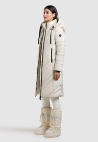 Lungo cappotto imbottito color crema con cuciture a forma di chevron, zip nere e cappuccio. Abbinato a stivali coordinati sopra il ginocchio per il freddo.
