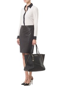 Femme portant un chemisier noir et blanc, une jupe en cuir noire, des talons hauts noirs et blancs, tenant un grand sac cabas noir avec des accents dorés.