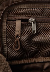 Brown mesh fabric with a zippered pocket and a hook. Includes silver zipper hardware and a brown strap detail. Textured surface visible.