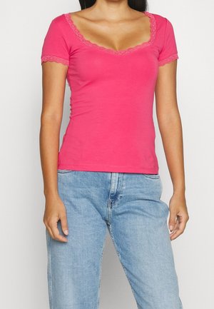 Kobieta ubrana w jasnoróżowy krótki top z koronkową lamówką i jasnoniebieskie dżinsy na jednolitym tle.