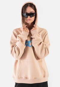 Beżowa, oversize bluza z kapturem z niebieskim logo, z przednimi kieszeniami i ribbrowymi mankietami. Model nosi czarne, kątowe okulary przeciwsłoneczne, a w tle widoczny jest gładki kolor.