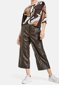 Kvinna iklädd en brun abstrakt mönstrad topp, mörkbruna vida cropped läderbyxor och vita sneakers, stående mot en vit bakgrund.