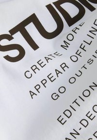 Bijela tkanina s velikim crnim natpisima, uključujući "STUDIO" i "STVARAJ VIŠE." Glatka struktura s mat finišem.