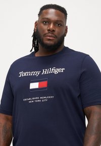 Marineblå t-skjorte laget av myk bomull, med en logo med et rektangulært design i rødt, hvitt og blått. Teksten leser "Tommy Hilfiger" og "Established MCMLXXXV New York City."