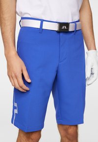 Niebieskie szorty golfowe o gładkiej strukturze, dopasowanym kroju i bocznych paskach. Połączone z białym paskiem i ręką noszącą rękawicę golfową.