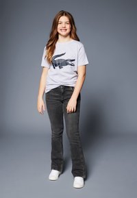 Jauna meitene smaida, stāv, vilkta pelēkā T-kreklā ar melnu krokodilā drukas rakstu, melnās džinsās un baltas kedās uz pelēka fona.