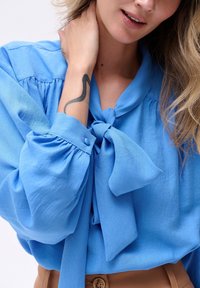 Jasnoniebieska bluzka z wiązaniem przy dekolcie, z bufiastymi rękawami i guzikami przy mankietach. Materiał wydaje się gładki i lekko błyszczący.