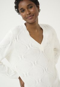 Hvid strikket sweater med V-udskæring og struktureret geometrisk mønster. Har lange ærmer og en afslappet pasform.