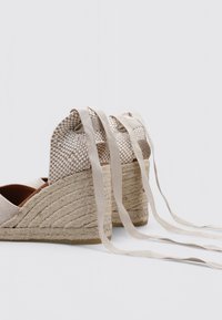 Cuñas de esparto beige con parte superior de tela trenzada, suelas de cuerda de yute y lazos de cinta en el tobillo. Punta redonda y detalle de superficie texturizada.