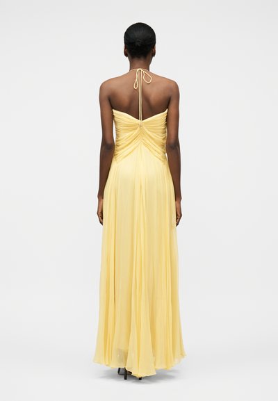 Žena v dlouhých, splývavých žlutých šatech na krk se shromážděným zády, stojící zády k nám na jednobarevném světlém pozadí.