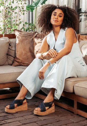 Blue denim wedge sandals with cork platform, crisscross strap design, and adjustable buckle. Model seated on a sofa, in a light outfit.