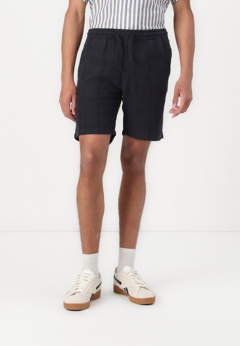 Zwarte shorts met een elastische tailleband, vallend tot halverwege de bovenbenen, gecombineerd met witte sneakers en lichte sokken, met een gladde stoftextuur.