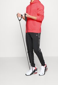 Sudadera roja con cordón, pantalones de chándal negros con logo blanco, y zapatillas blancas con acentos rojos, sosteniendo bandas de resistencia.