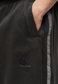 Χέρι μέσα στην τσέπη μαύρου φόρμας με ανακλαστική πλευρική ρίγα και κεντημένο λογότυπο "favela" κοντά στην τσέπη.