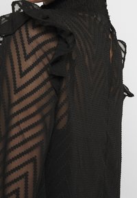 Blouse noire transparente avec un motif en zigzag texturé, détail volanté aux épaules et fermeture à boutons au dos. Tissu léger.