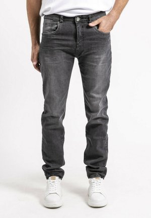 Homme portant un jean slim gris foncé, main dans la poche et baskets blanches, debout devant un fond blanc uni.