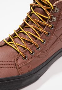 Hnědé kožené vysoké tenisky se žlutými a hnědými tkaničkami, stříbrnými očky a černou gumovou podrážkou s texturovanými detaily.