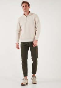 Világos bézs, cipzáros pulóver, bordás ujjak és szegélyekkel, sötétzöld, slim-fit nadrággal és bézs sportcipővel, barna kiegészítésekkel párosítva.