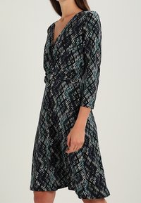 Knä-lång klänning i marinblått med ett geometriskt mönster i turkos och vitt. Har en V-ringning och trekvartsärmar, tillverkad av mjuk tyg.