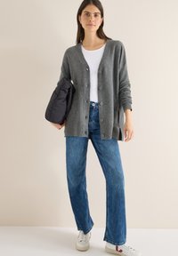 Cardigan grigio a maglia con scollo a V e bottoni, indossato sopra una t-shirt bianca e jeans blu. Tiene una borsa nera, in piedi su uno sfondo chiaro.