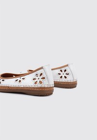 Zapatos planos blancos con recortes florales, acentos marrones y una suela texturizada. Presentan un detalle de costura alrededor de los bordes.