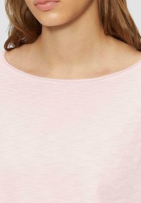 Haut en tissu doux, rose pâle, avec un décolleté large. Montre une texture lisse et un design décontracté, avec des détails au niveau du col et des épaules.