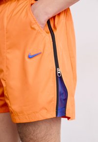 Oransje Nike-shorts med en sidelomme med glidelås, utstyrt med blå mesh-fôr og en merkede logo i blått. Laget av lett, glatt stoff.