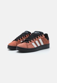 Suede sneakers in rust with black accents, featuring three white stripes, black laces, and a textured rubber sole.