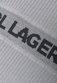Tessuto in maglia argento con una banda orizzontale nera con logo, caratterizzato da una texture a coste e un leggero luccichio.