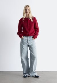 Rote, kuschelige Strickjacke mit Knöpfen, kombiniert mit hoch geschnittenen hellblauen weiten Jeans und dunklen Schuhen mit weißen Streifen.