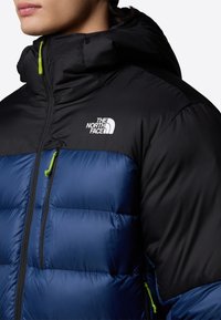 Czarnie i niebieska kurtka puchowa z zamkiem przy szyi, zielonym uchwytem zamka oraz logo na górnej lewej stronie. Teksturowana tkanina z pikowanymi sectionami.