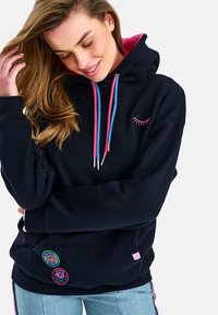 Mladá žena v černé mikině s kapucí, s růžovými a modrými šňůrkami, výšivkou řas na hrudi a květinovými nášivkami u kapsy.