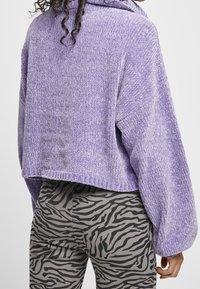 Lila, kurz geschnittenes Sweatshirt mit einem strukturierten, gerippten Muster und einer Kapuze. Kombiniert mit grauen Leggings, die ein schwarzes Zebra-Streifenmuster aufweisen.