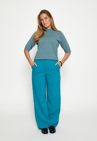 Trumpa rankovėmis, danguje mėlyna palaidinė su aukštu kaklu, derinama su plačiomis mėlynomis kelnėmis. Modelis pozuoja su rankomis kišenėse, avintis juodus batus.