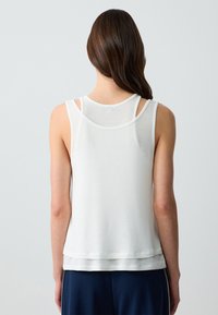 Top bianco senza maniche con un design a strati, caratterizzato da una texture liscia e sottili accenti cuciti. La vista posteriore mostra spalline incrociate.