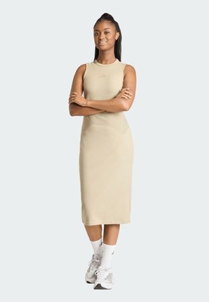 Giovane donna in piedi con le braccia incrociate, indossa un vestito midi beige senza maniche, calzini bianchi e scarpe da ginnastica bianche su uno sfondo neutro.