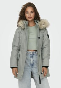 Parka grise avec une capuche bordée de fourrure, fermeture éclair et bouton, taille ajustable et accents noirs contrastants sur les manches et les poches.