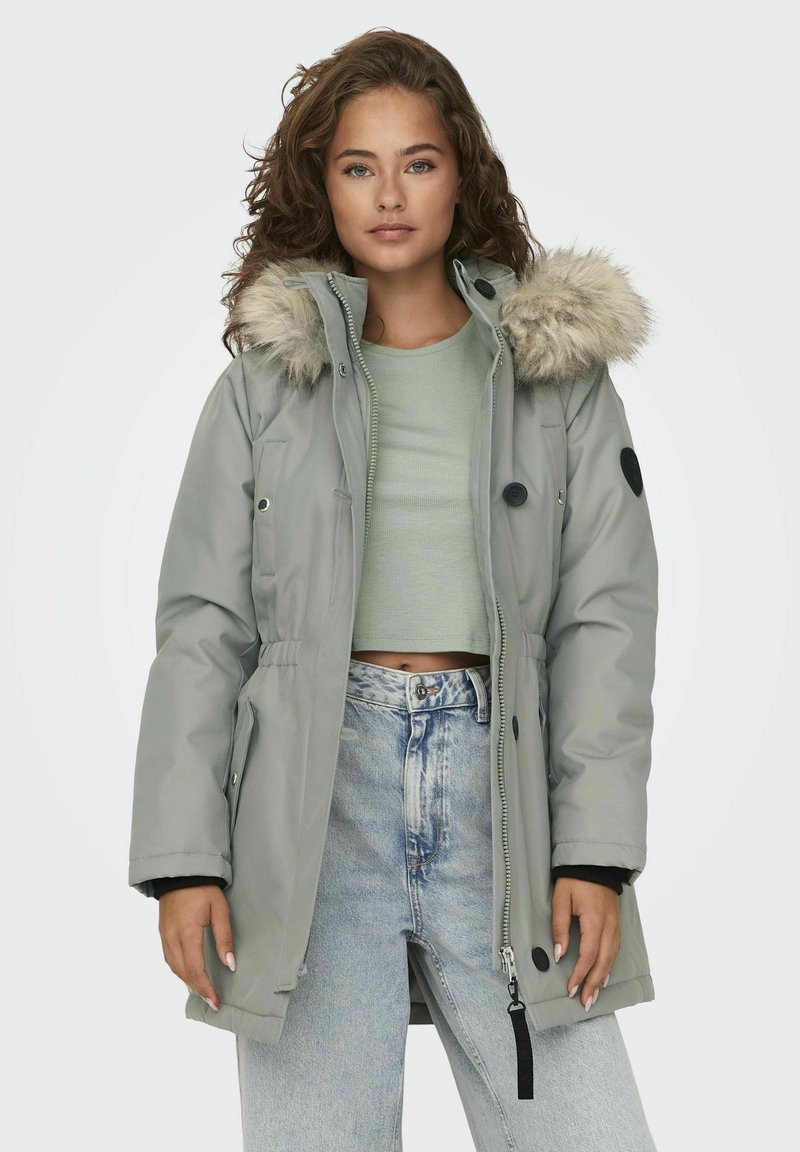 Parka grise avec une capuche bordée de fourrure, fermeture éclair et bouton, taille ajustable et accents noirs contrastants sur les manches et les poches.