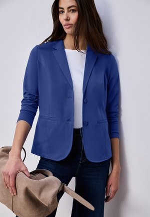 Femme portant un blazer bleu sur une chemise blanche et un jean foncé, tenant un grand sac en daim beige avec une bandoulière.